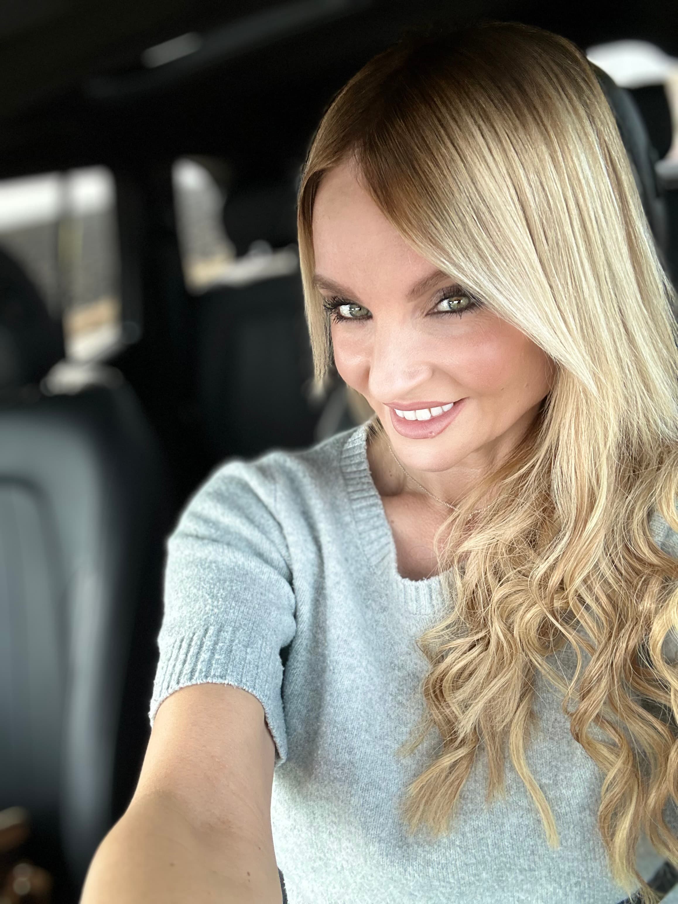 Smiling blonde woman with long wavy hair wearing a grey sweater in a car.