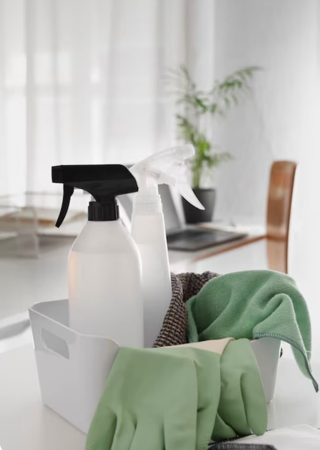 White bin holding spray bottles, green cleaning cloth, and rubber gloves in a bright room.